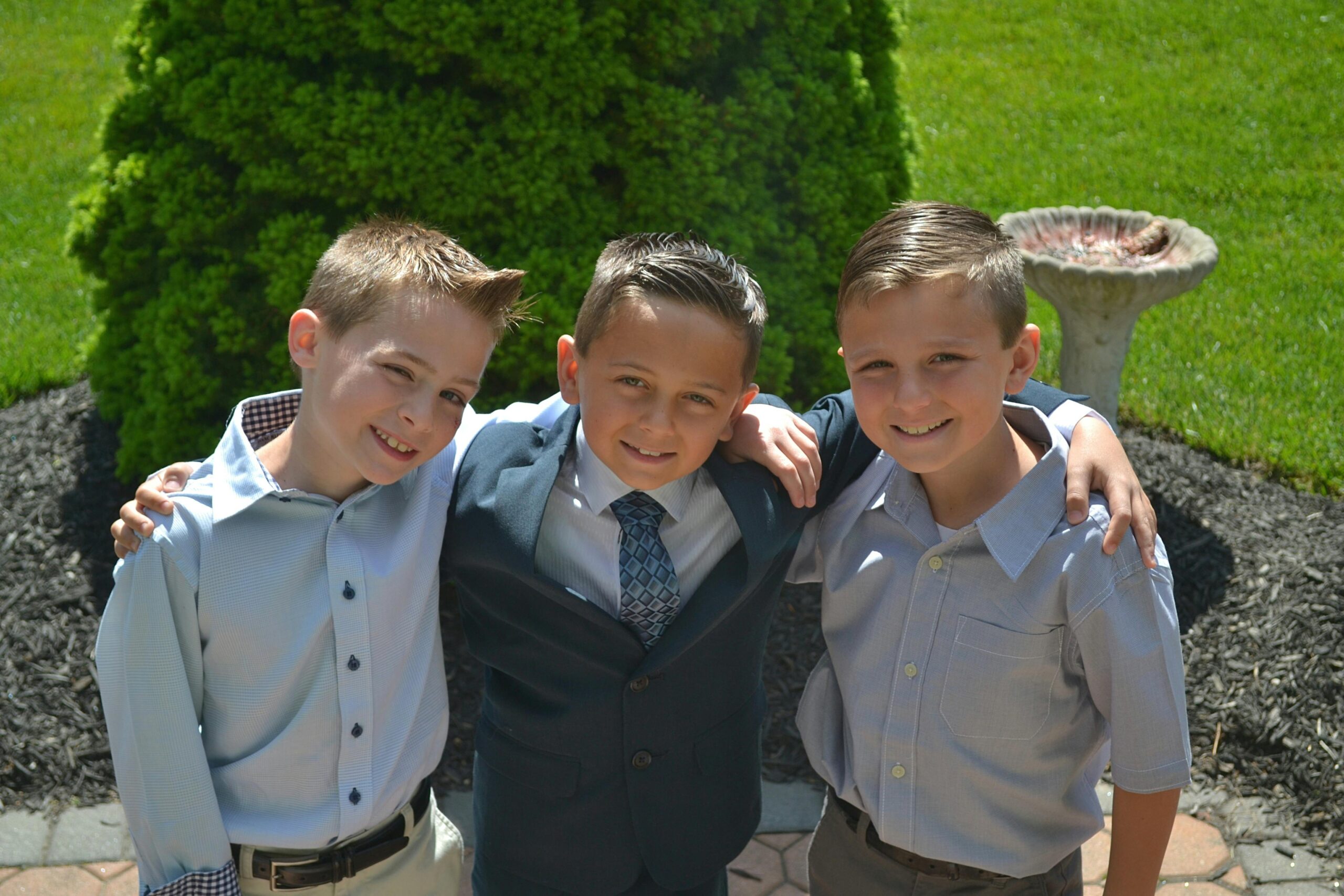 Three boys in formal shirts smiling together outdoors in a sunny garden.