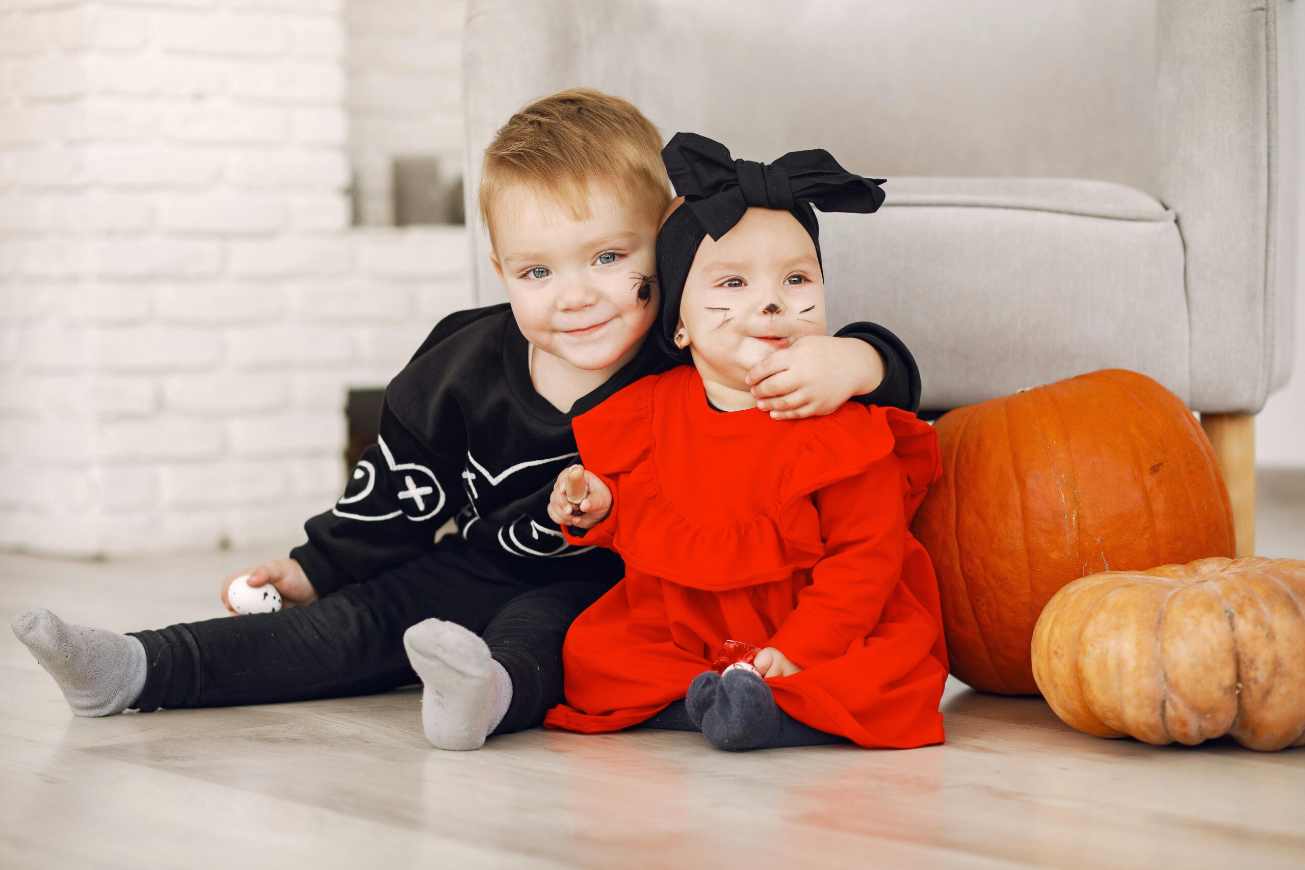 Two cute toddlers in Halloween costumes sitting indoors with pumpkins, ready for fall festivities.