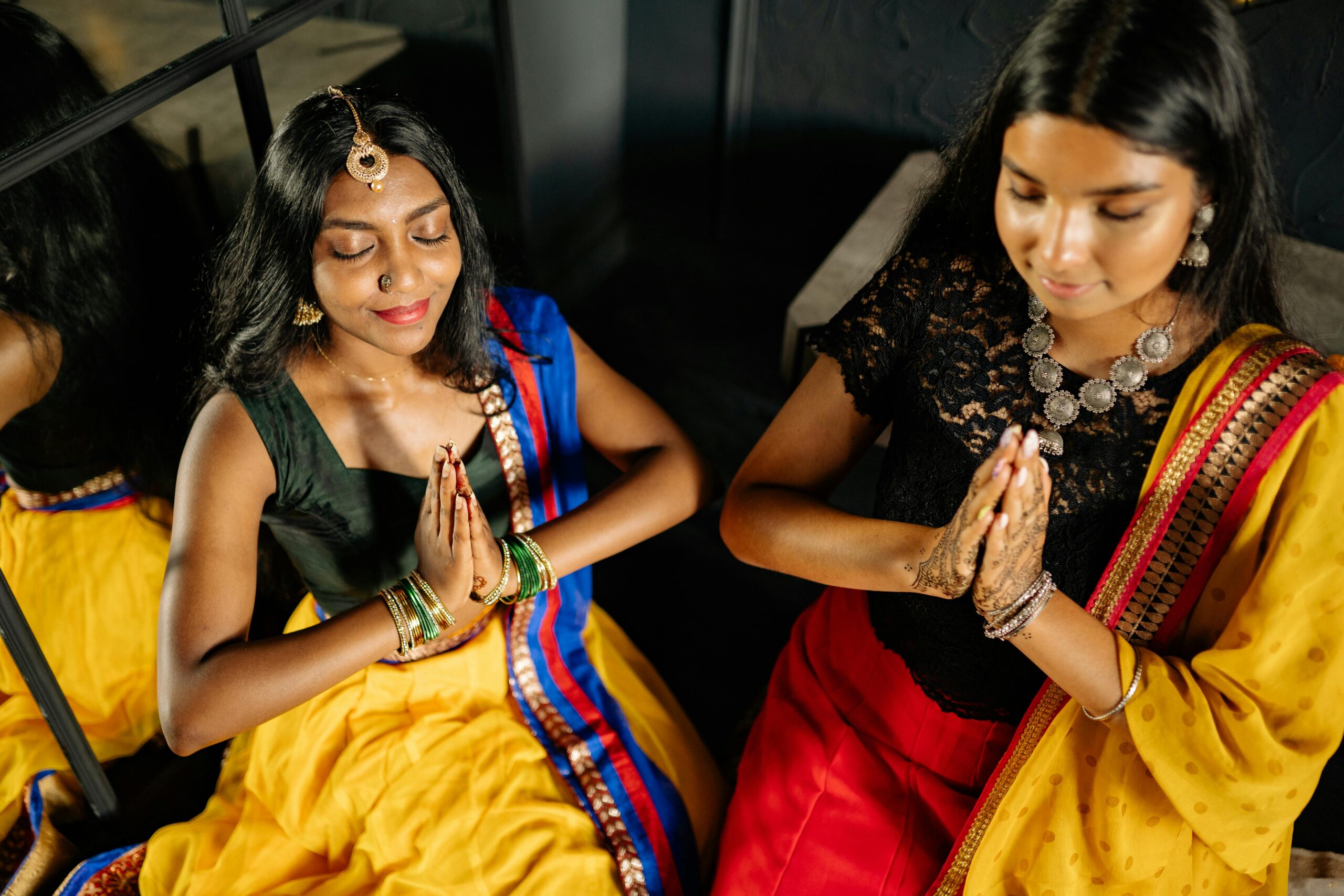 Two women in traditional Indian dresses meditate with praying hands indoors.